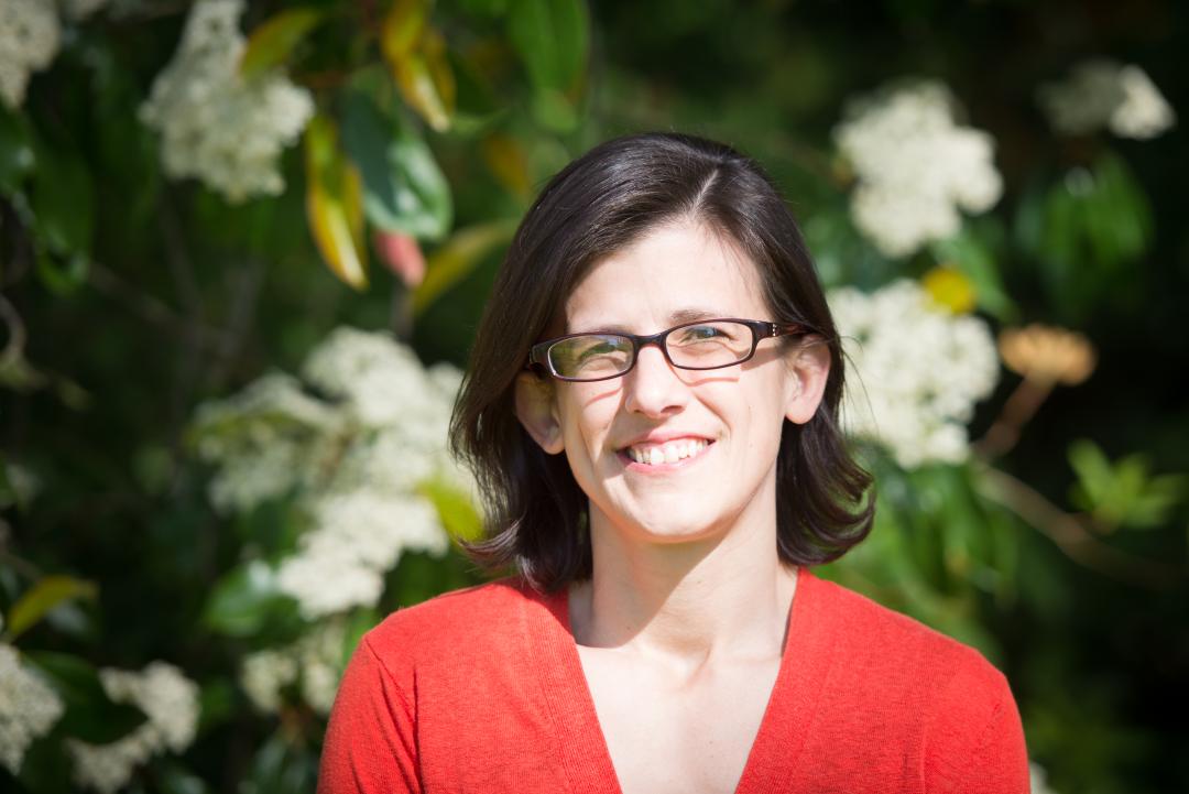 A woman in a red shirt with dark hair stands in front of a green leafy background with white flowers.