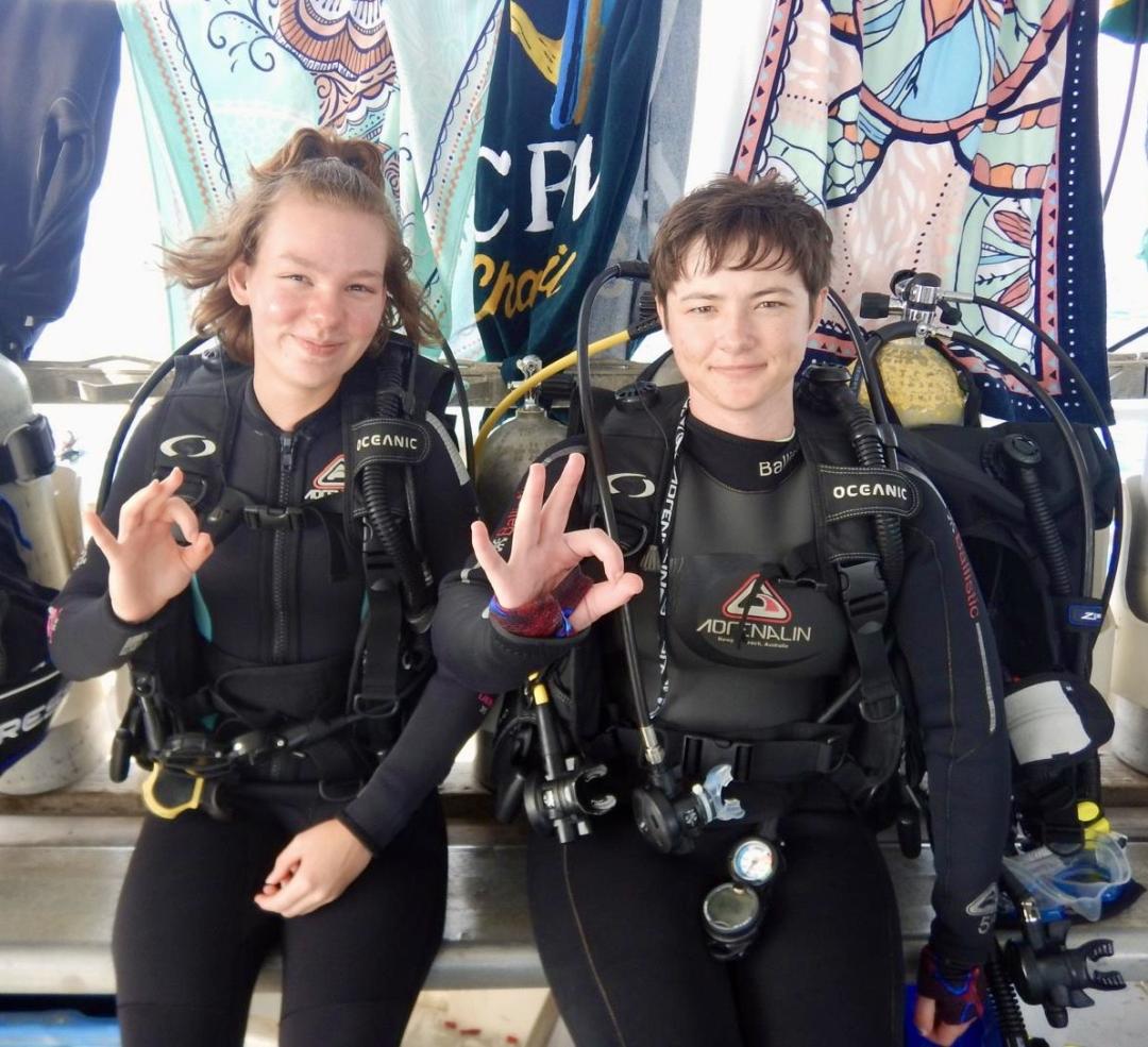 Two scuba divers hold up the ok symbol while sitting on a boat.