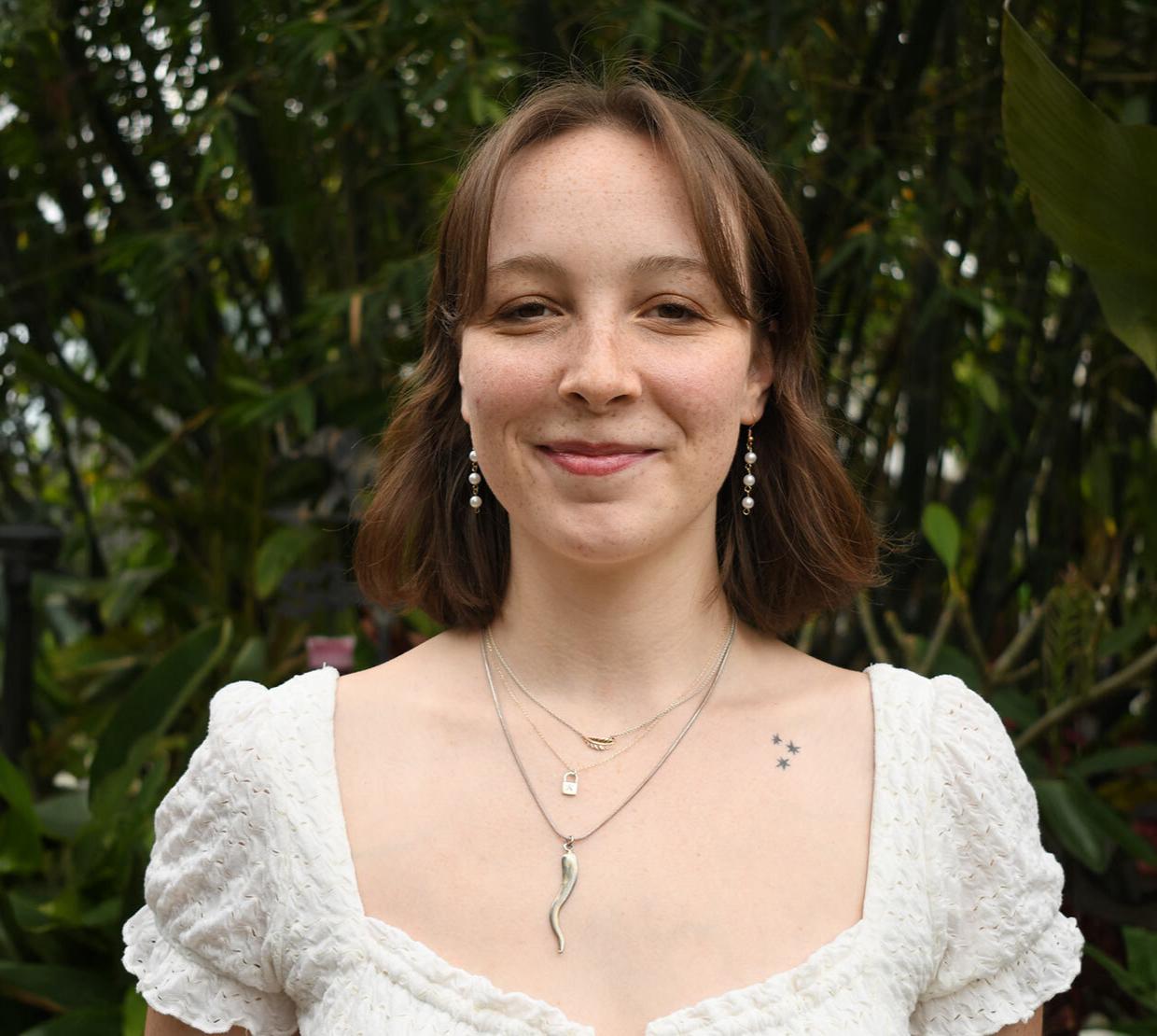 A woman in a white blouse stands in front of greenery. 