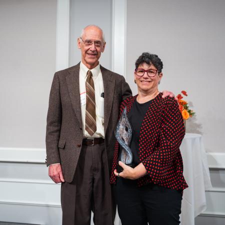 A man in a brown suit stands next to a woman wearing a red and black dress jacket. The woman is holing a glass award.