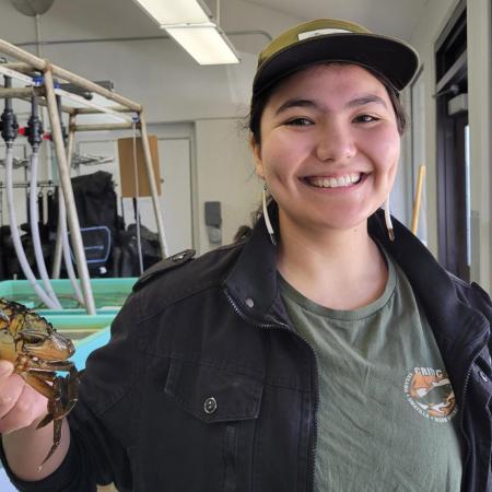 Ingram standing the Hatfield Marine Science Center wet lab, holds up a crab. She wears a CRITFIC shirt, baseball cap and beaded earrings.