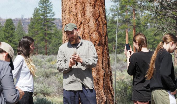 A man holds a bird and shows student. Background is trees and grass.