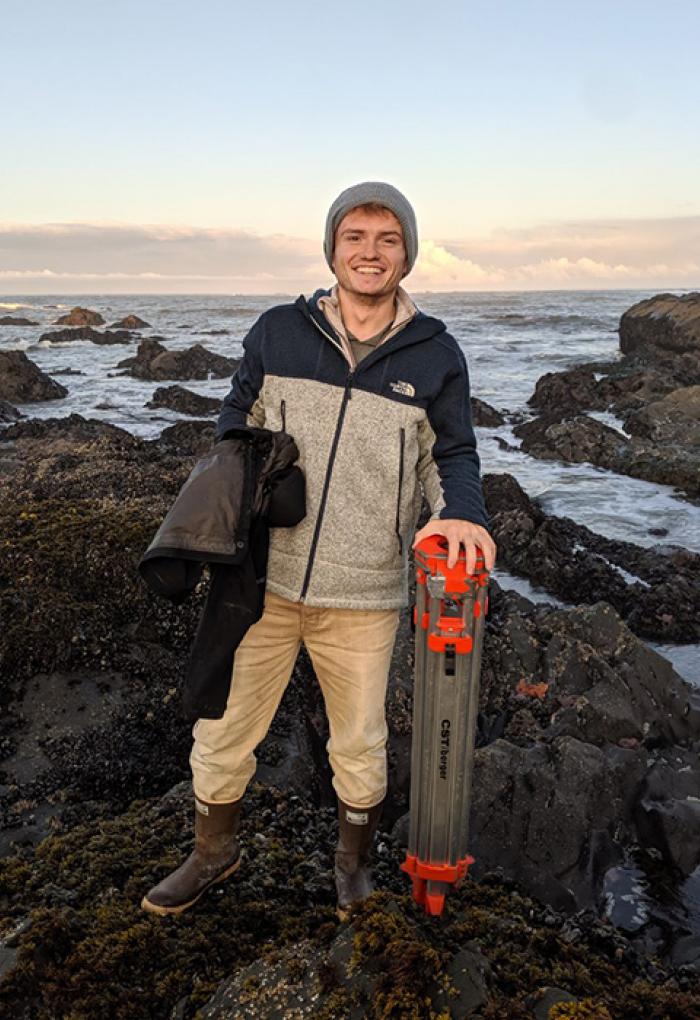Kristofer Bauer standing on rocks on ocean shore with research equipment