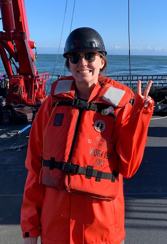Woman in orange jacket and life vest giving the peace sign aboard a ship at sea.